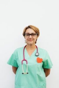 Portrait of a confident female doctor in green scrubs with a stethoscope and eyeglasses against a white background.